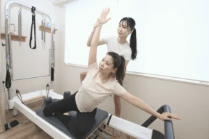 女性トレーナーがお客様の腕を支えながら、リフォーマーの上で体側を伸ばすマシンピラティスのパーソナルレッスン