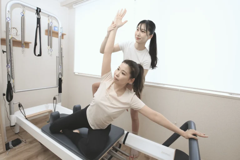 女性トレーナーがお客様の腕を支えながら、リフォーマーの上で体側を伸ばすマシンピラティスのパーソナルレッスン